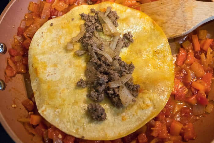 Dip the tortilla in the tomato, onion, and red bell pepper mixture, then add a layer of the ground beef mixture on top of the tortilla.