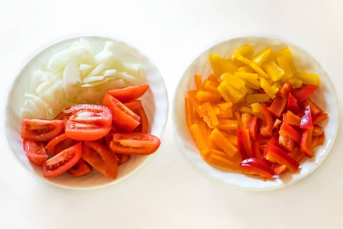 Sliced onions, yellow, red, and orange bell peppers, and tomatoes are sliced for the pepper steak recipe.