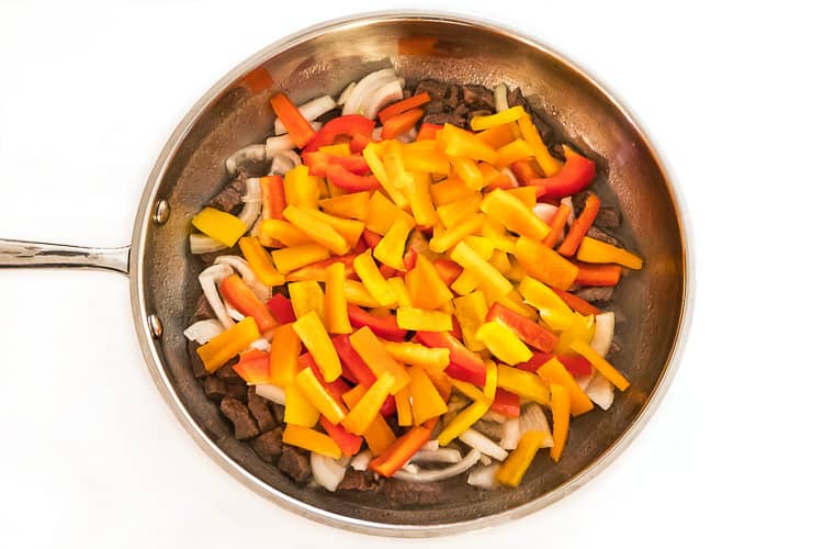 Steak, onions, yellow, red and orange bell peppers in a frying pan.
