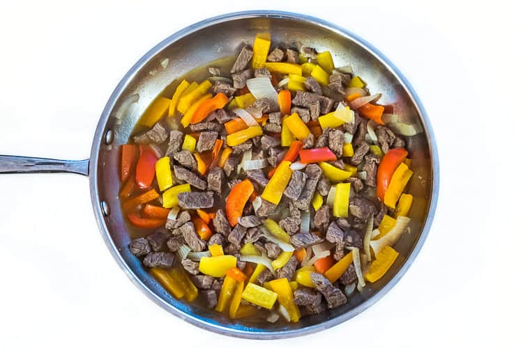 Steak, onions, yellow, red, and orange bell peppers in a frying pan.